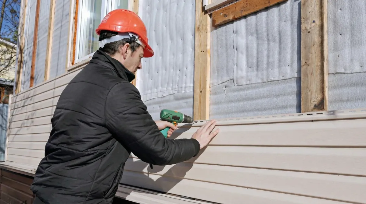 Siding replacement professional using a power drill to install new tan vinyl siding on a home exterior in Bloomington, IN